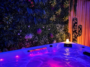 A hot tub with a waterfall feature surrounded by lush greenery and a wooden wall.