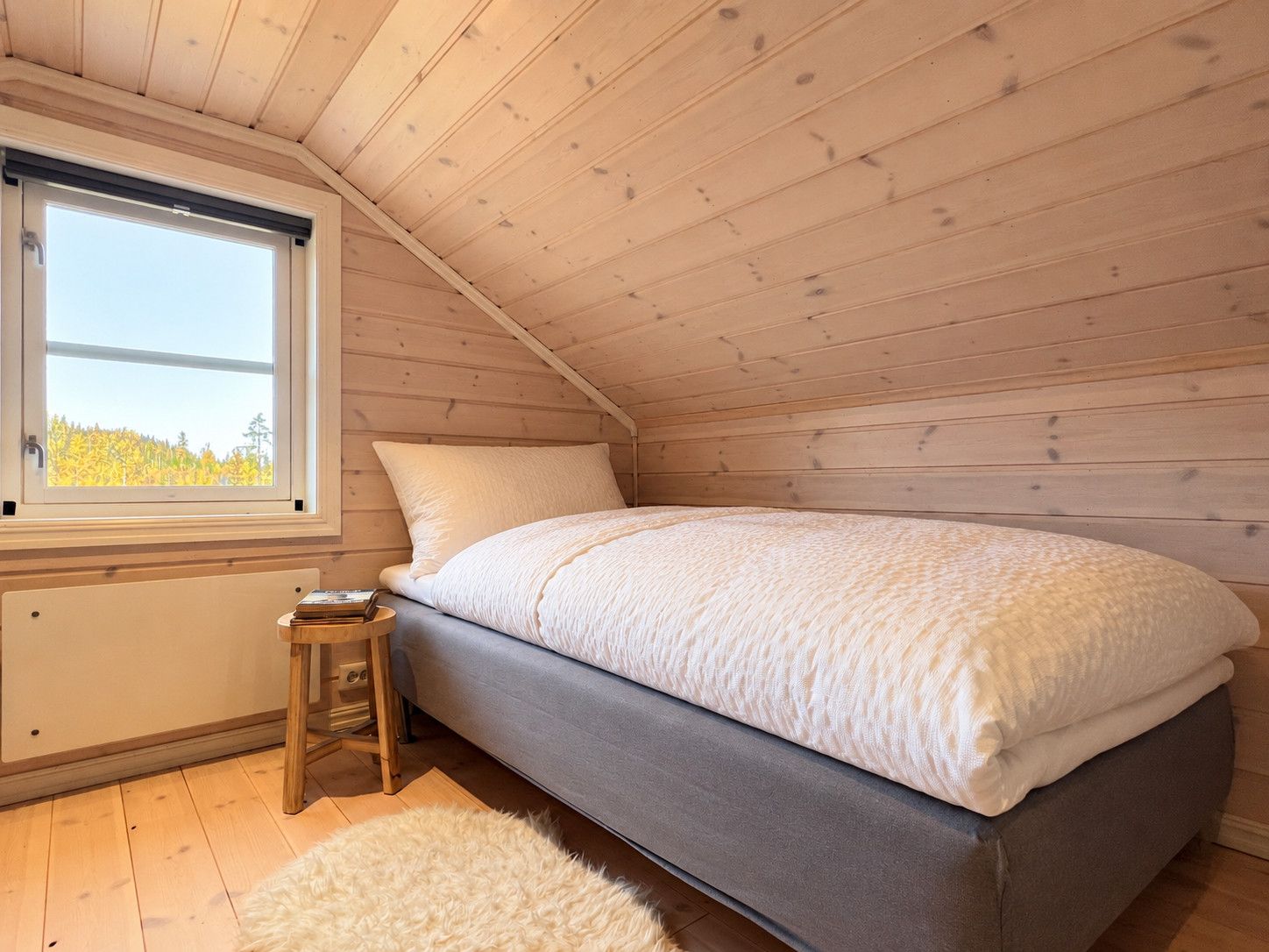 A bedroom with a single bed, a small wooden stool, and a window with a view of trees.