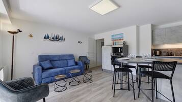 Een moderne woonkamer met een blauwe bank, een eethoek met een ronde tafel, en een keuken met een inbouwoven.