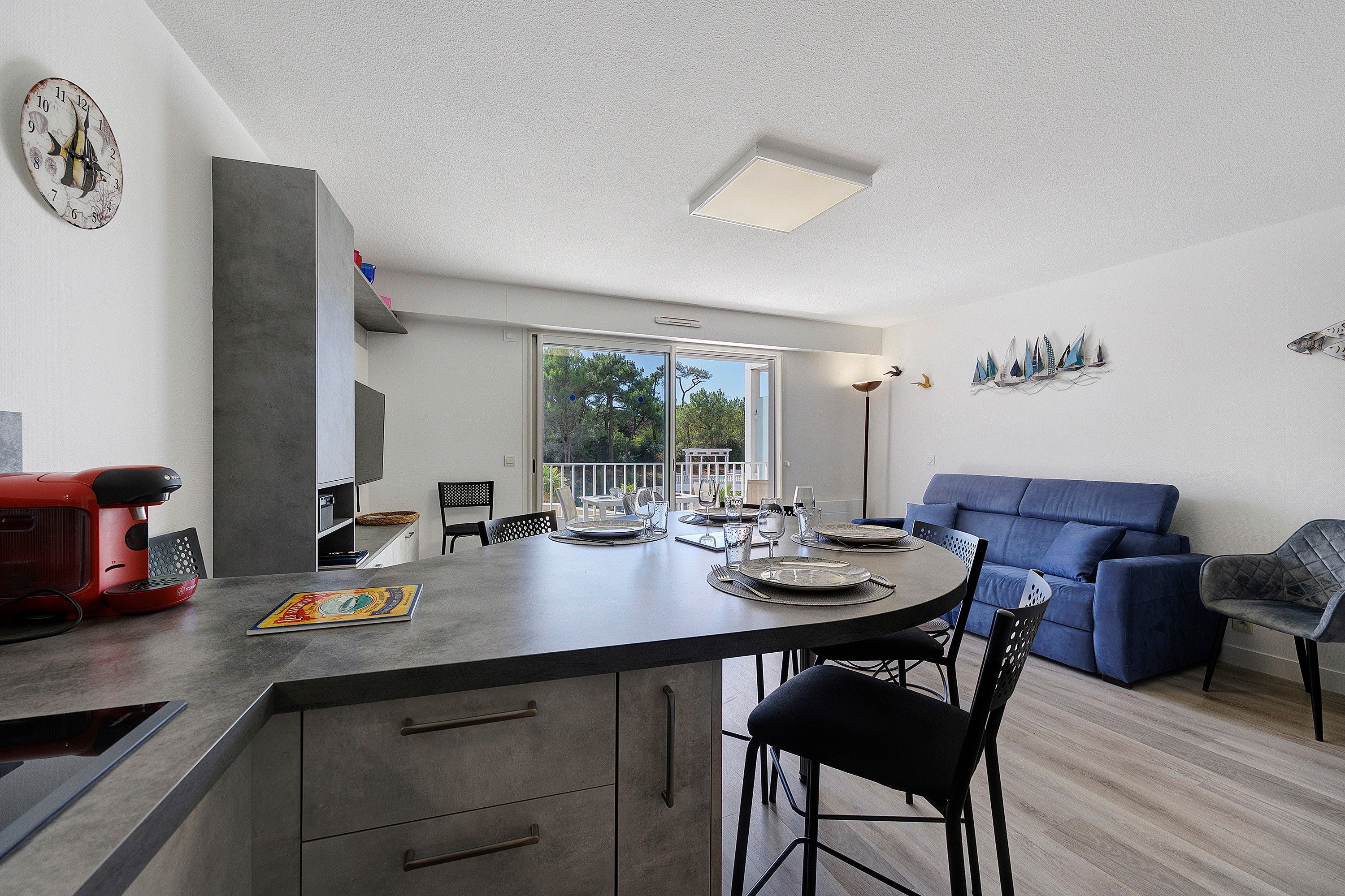 A modern kitchen with a central island, dining area with chairs, a blue sofa, and a wall-mounted clock.