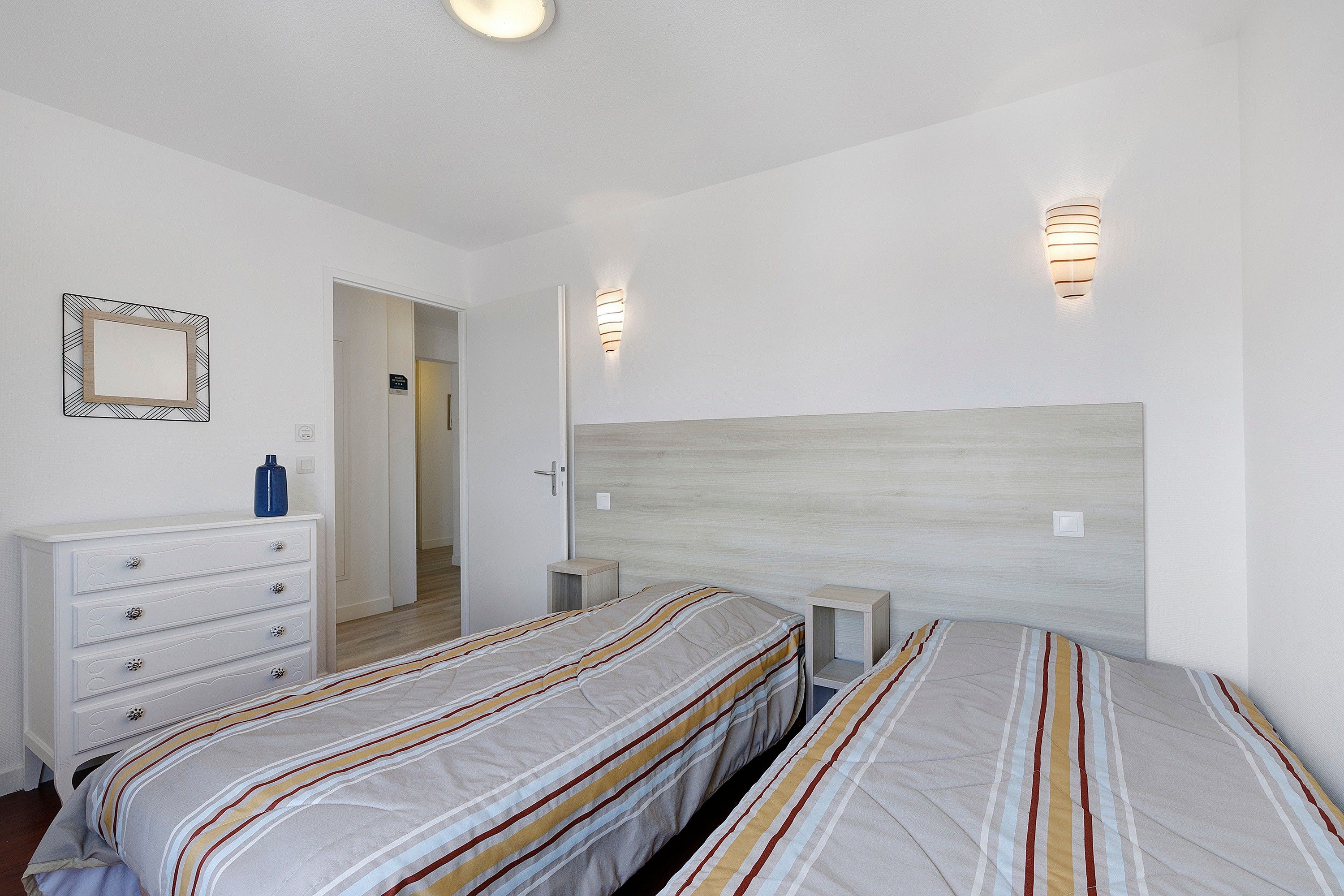 Une chambre avec deux lits, une commode, un miroir et deux lampes fixées au mur.