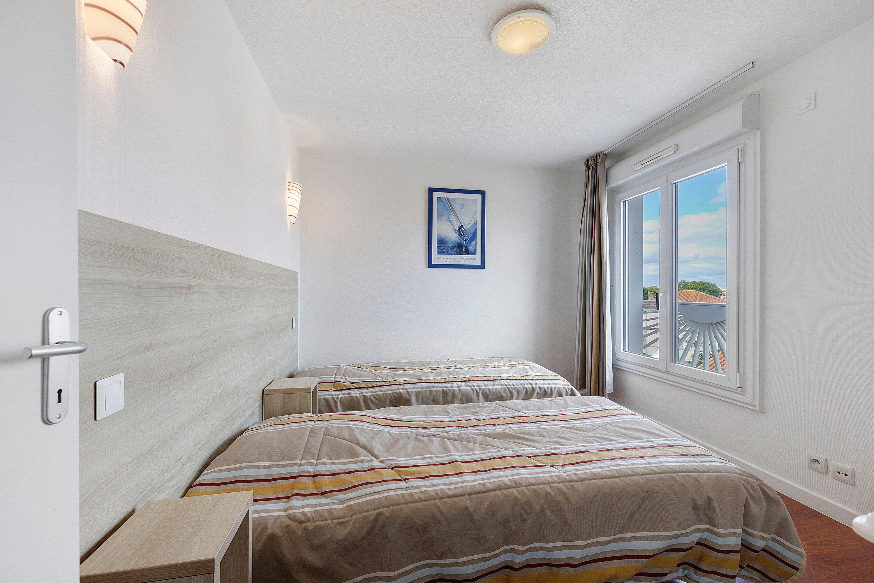 Une chambre d’hôtel avec deux lits, une fenêtre donnant sur un paysage agréable, un tableau accroché au mur et une porte dotée d’une poignée.