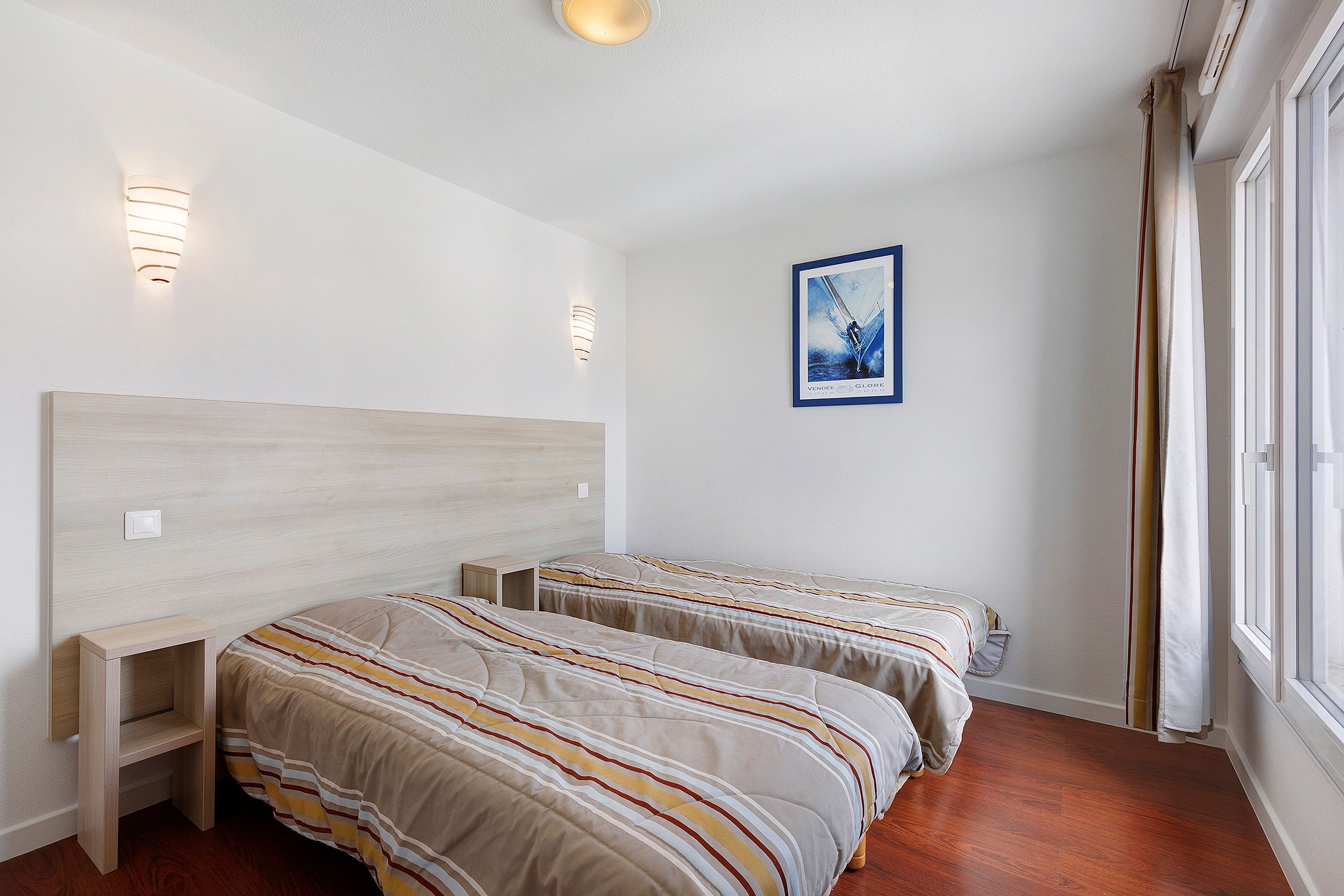 Une chambre d’hôtel avec deux lits, une table de chevet, un tableau encadré au mur et une fenêtre avec des rideaux.