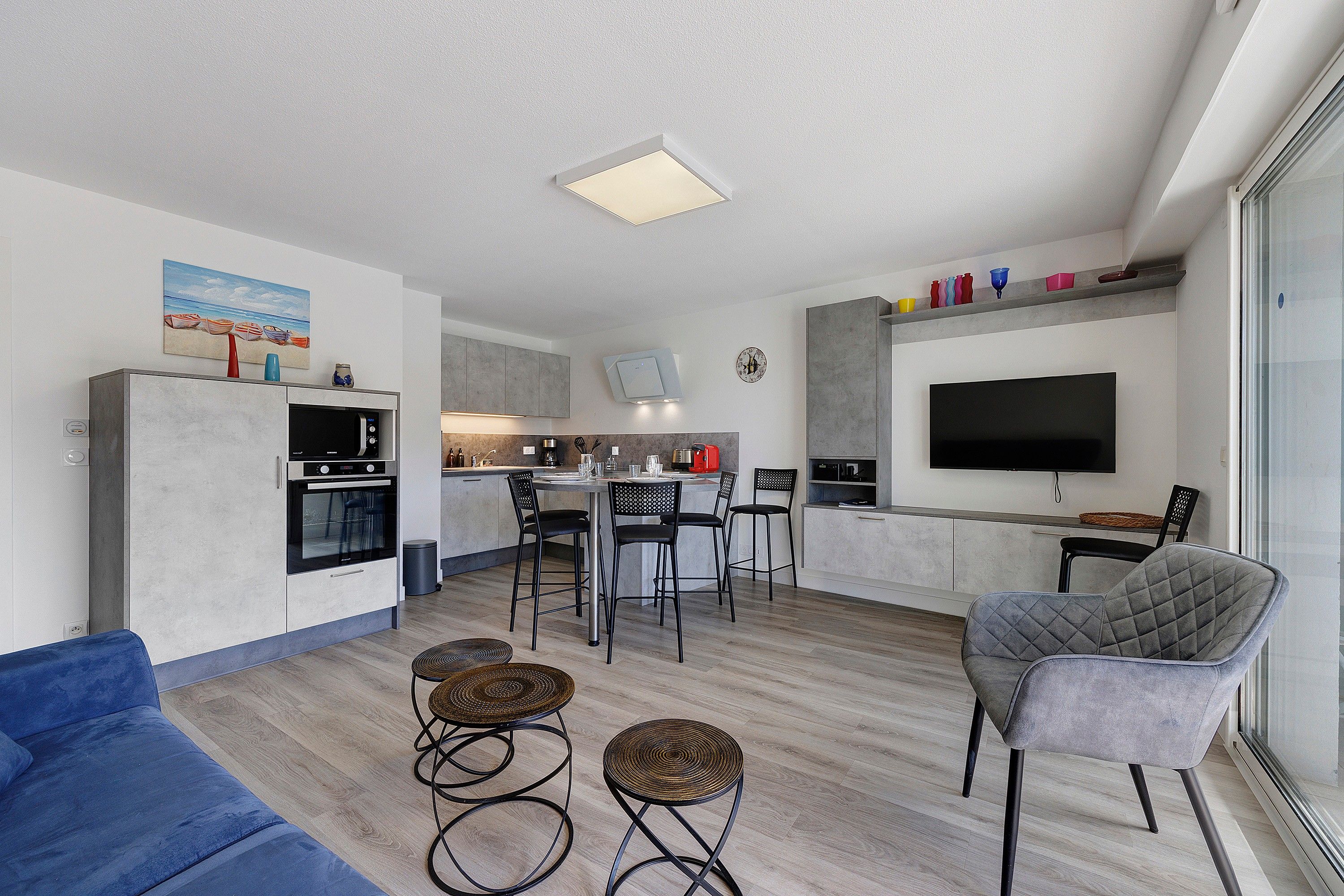 Een moderne woonkamer met een blauwe bank, twee ronde rieten krukjes, een grijze fauteuil en een flatscreen televisie aan de muur.