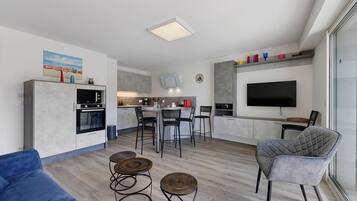 Een moderne woonkamer met een blauwe bank, twee ronde rieten krukjes, een grijze fauteuil en een flatscreen televisie aan de muur.