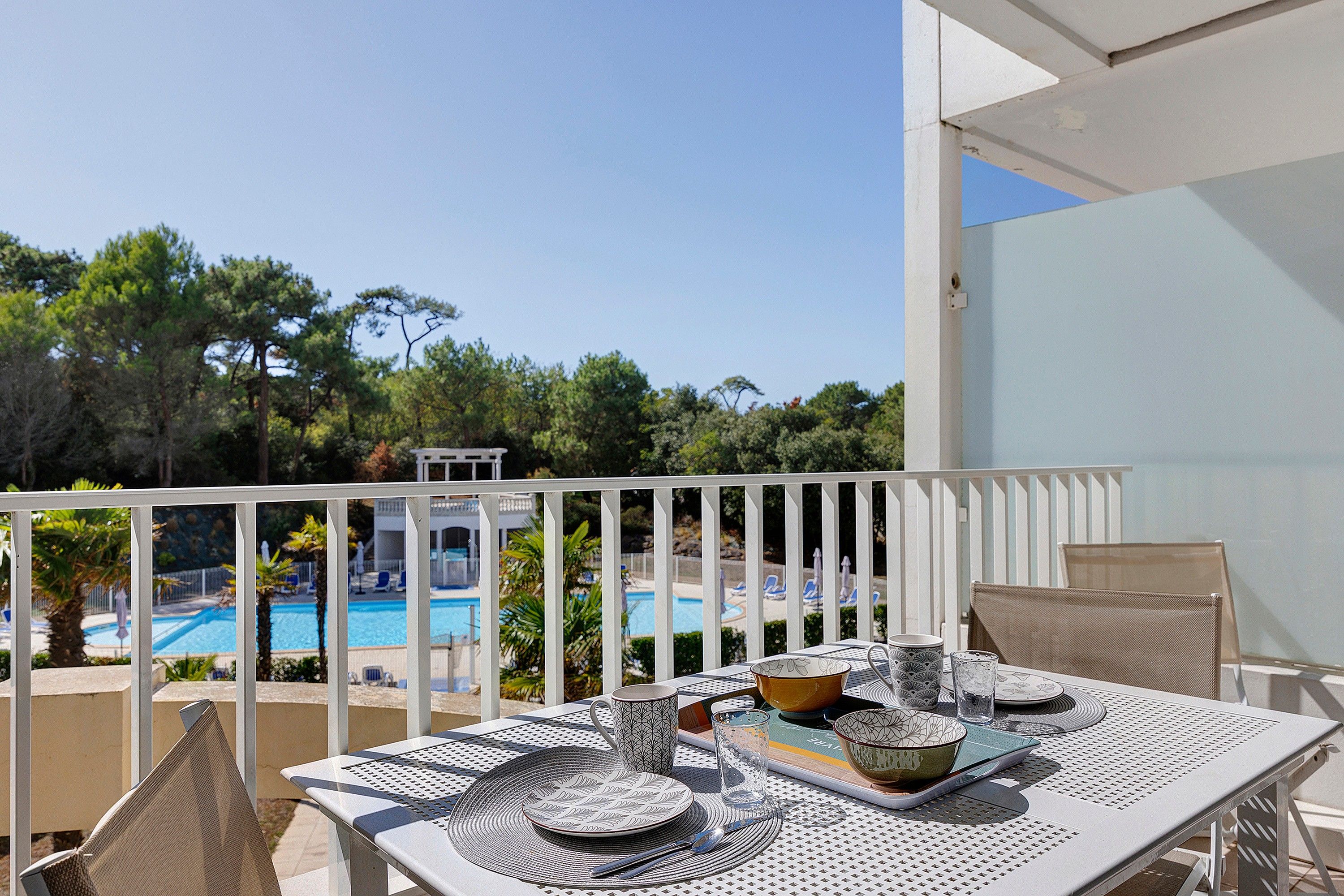 Un balcon avec une table dressée pour le dîner, donnant sur une piscine et des arbres.