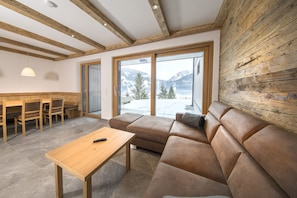 Ein modernes Wohnzimmer mit Holzboden, einer Couch, einem Tisch und Blick auf verschneite Berge durch große Fenster.