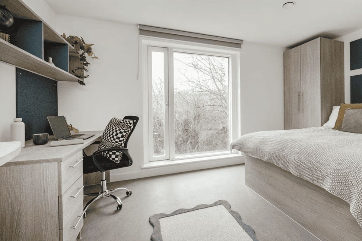 A bedroom with a bed, a desk, a chair, a window, and a wardrobe.