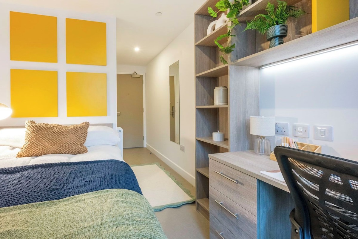 A hotel room with a bed, bedside table, lamp, and a desk with a chair.