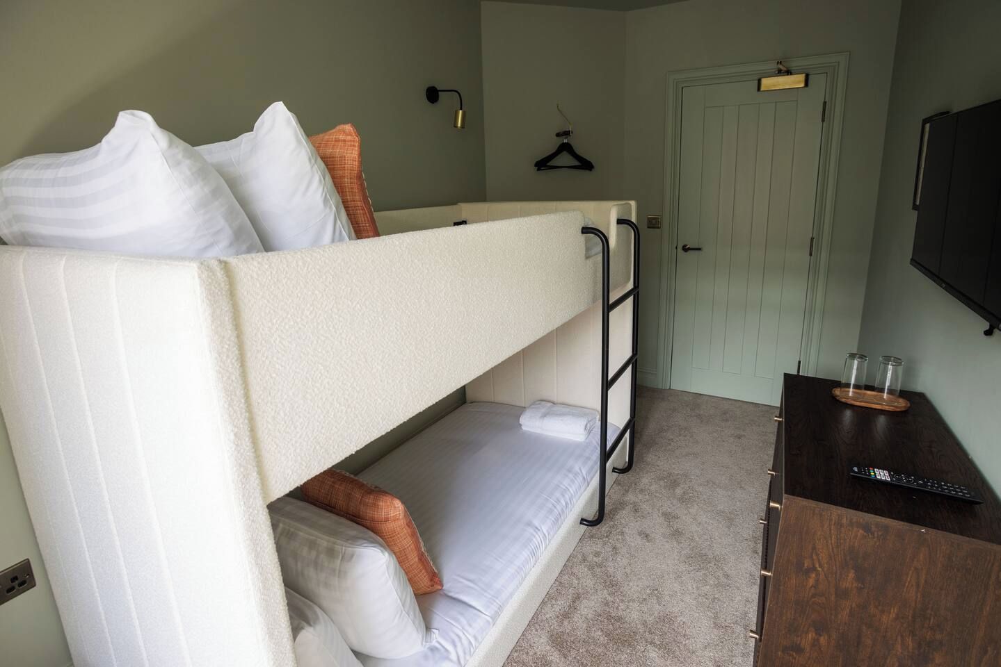 A bunk bed with pillows, a bedside table with a remote control, a coat rack, and a door in a room with light green walls.