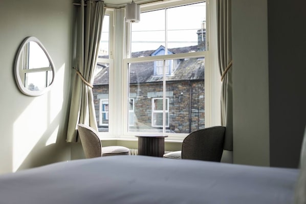 A room with a bed, two chairs, a round mirror, and a window with curtains.