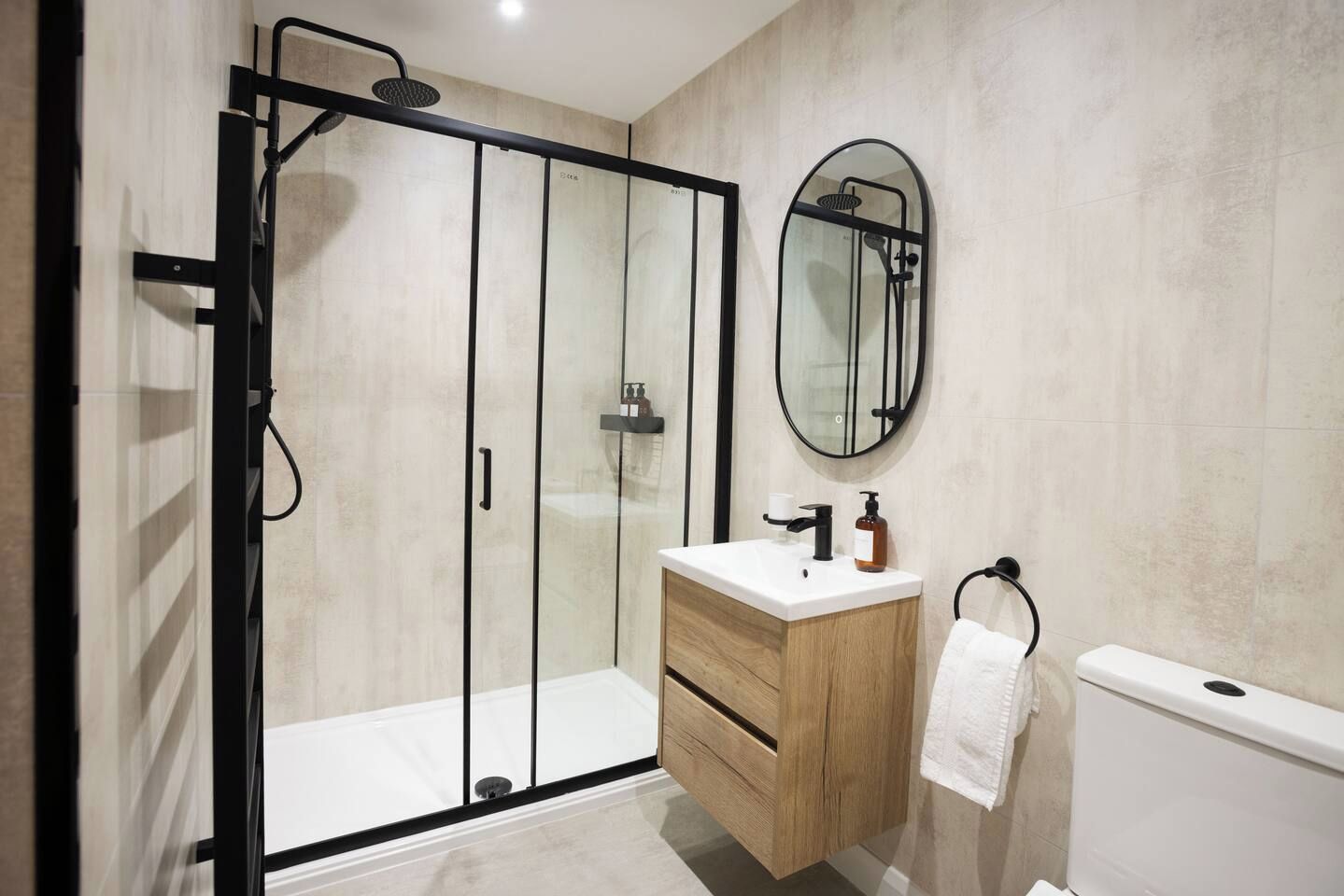 Un baño moderno con ducha envuelta en vidrio, tocador de madera, espejo redondo y un inodoro blanco.