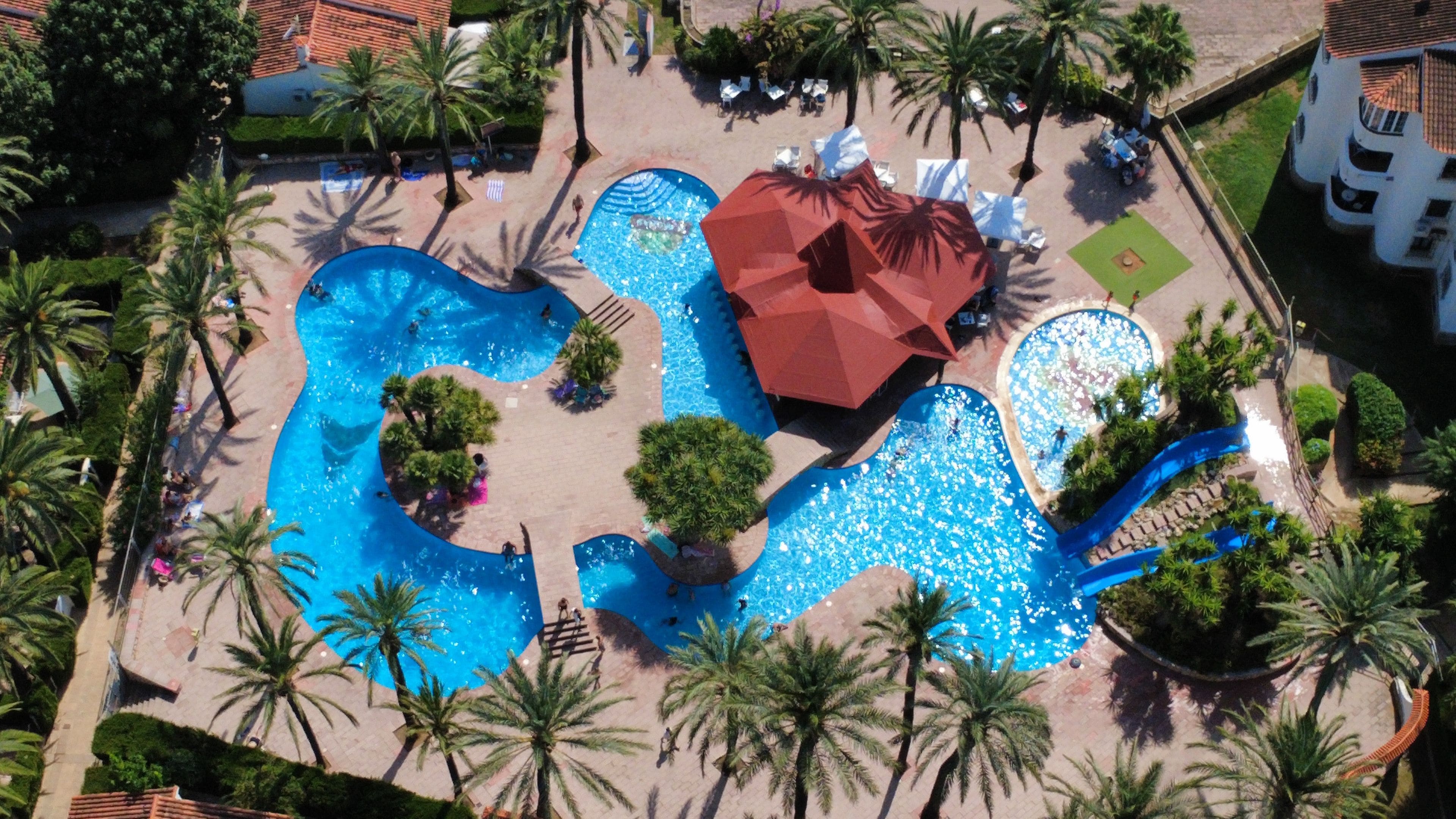 Vista aérea de un complejo turístico con varias piscinas, palmeras y una estructura de techo rojo.
