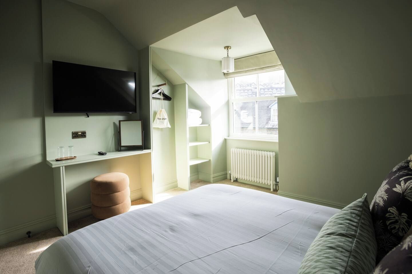 A bedroom with a bed, a desk, a TV mounted on the wall, a radiator, and a window with a view.