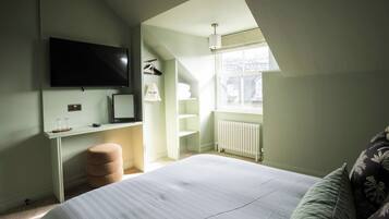 A bedroom with a bed, a desk, a TV mounted on the wall, a radiator, and a window with a view.