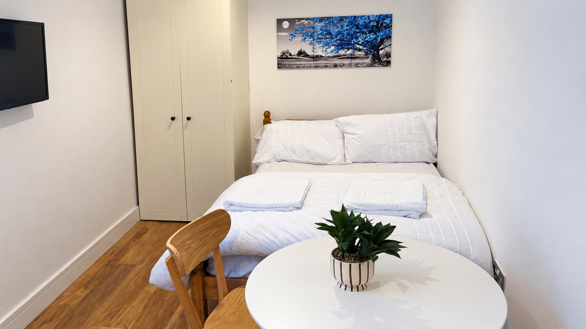 Une chambre à coucher avec un lit, une table de chevet ornée d’une plante, une chaise en bois, une armoire et un tableau accroché au mur.