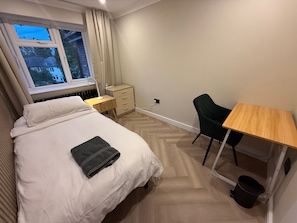 A hotel room with a bed, a desk, a chair, a radiator, and a window with curtains. - 5 Bed Home - Contractors & Professionals - Wifi (Luton)