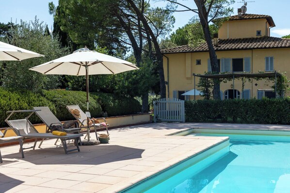 Ein Pool mit Liegestühlen und Sonnenschirmen, umgeben von einer Hecke und einem Gebäude mit Ziegeldach.