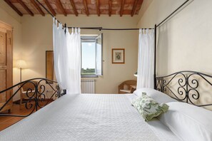 Ein Schlafzimmer mit Bett, Vorhängen, einem Fenster, einem Sessel und einer Lampe.