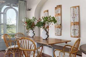 Uma sala de jantar com mesa de madeira e cadeiras de vime, dois vasos com plantas e decoração rústica nas paredes.