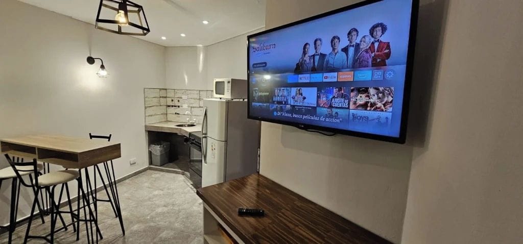 Una sala moderna con televisor de pantalla plana montado en la pared, una mesa de comedor de madera con sillas y una cocina con microondas y armarios.