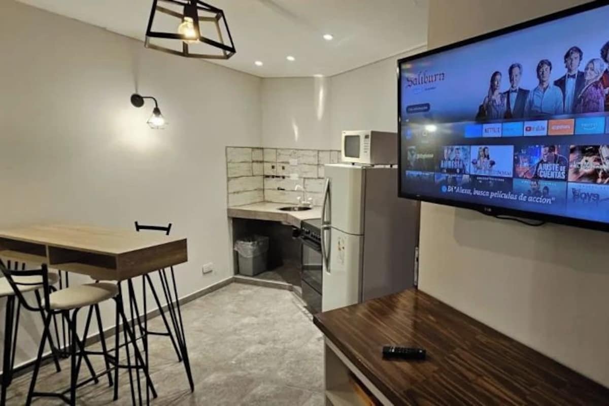 A modern living room with a flat-screen TV mounted on the wall, a wooden dining table with chairs, and a kitchen area with a microwave and cabinets.