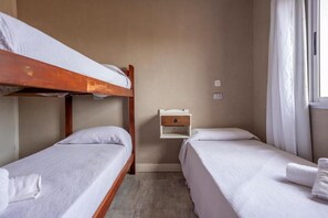 Habitació amb dos llits individuals, una lliterà i una petita taula de nit amb calaix, i una finestra amb cortines.