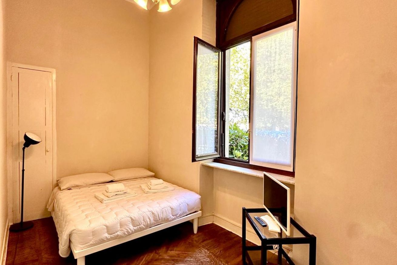 Une chambre à coucher avec un lit, une fenêtre donnant sur un paysage verdoyant, une table de chevet et une lampe sur pied.