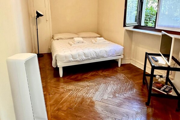 A bedroom with a bed, a standing lamp, a table with a laptop, and a window with a view of greenery. - Stunning apartment for two, just steps from Parco del Valentino – Turin. (Turin)
