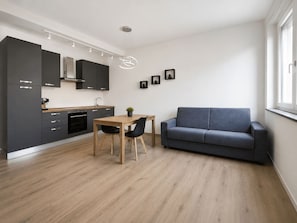 A modern kitchen with dark cabinets, a wooden dining table, and a grey sofa.
