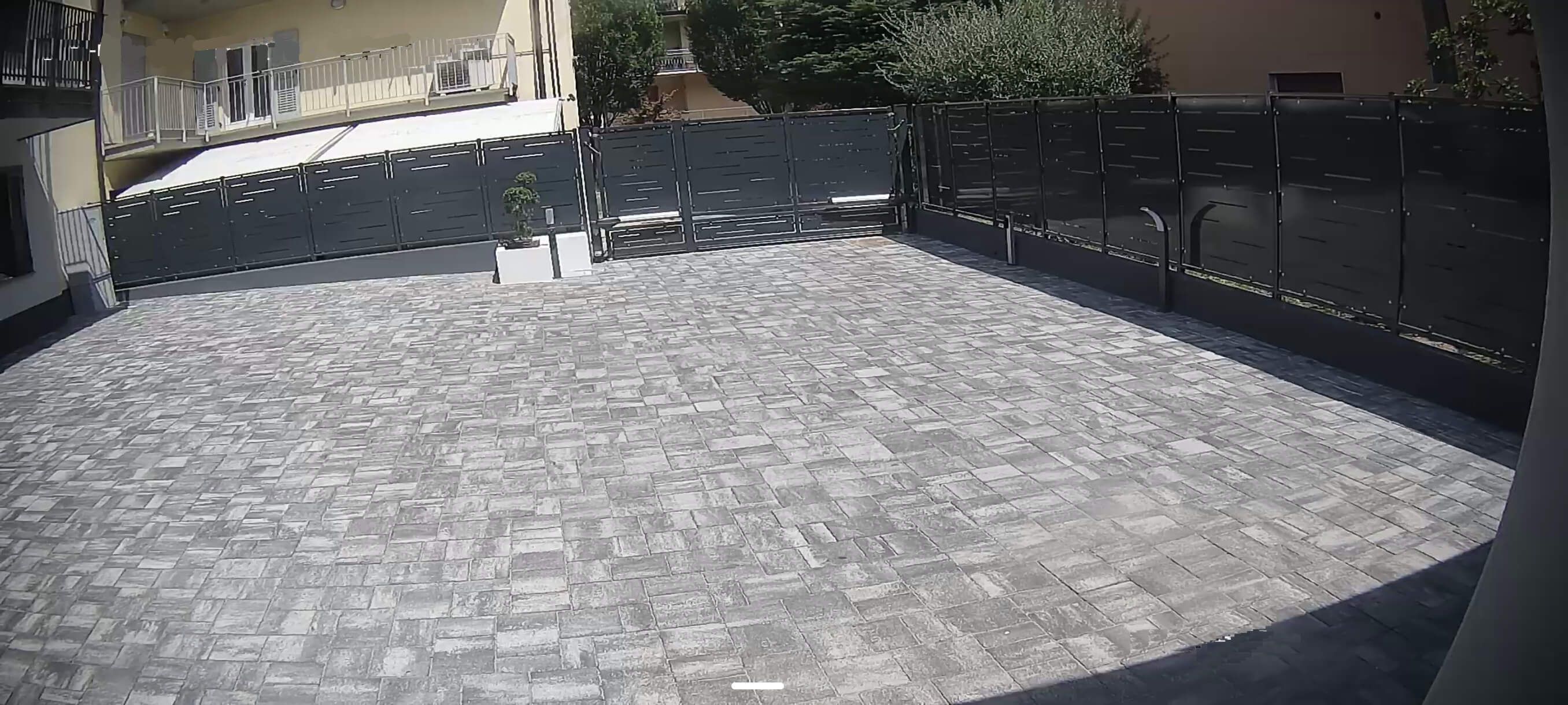 A paved outdoor area with a black metal fence, a small white planter, and a building in the background.