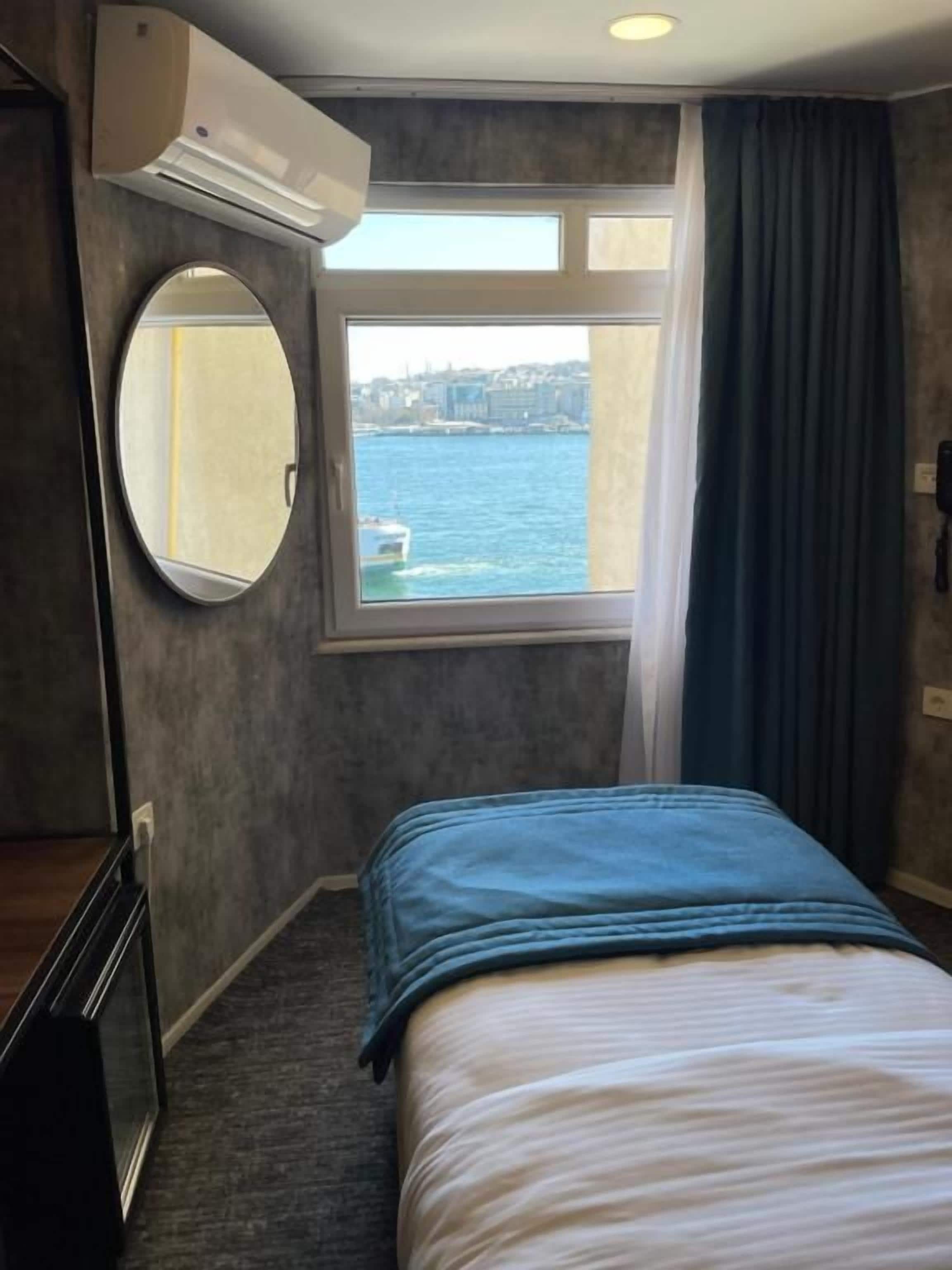 Habitación de hotel con cama, ventana con vista al agua, espejo redondo y unidad de aire acondicionado.