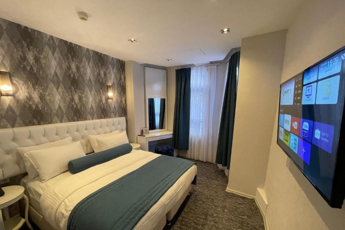 A hotel room with a bed, a desk, a TV, and a wall-mounted control panel.