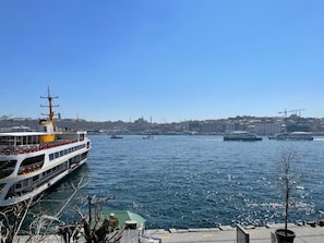 A river with a large ferry and several smaller boats, a clear sky, and a cityscape in the background. - Weingart Port Seaside Hotel (Istanbul)