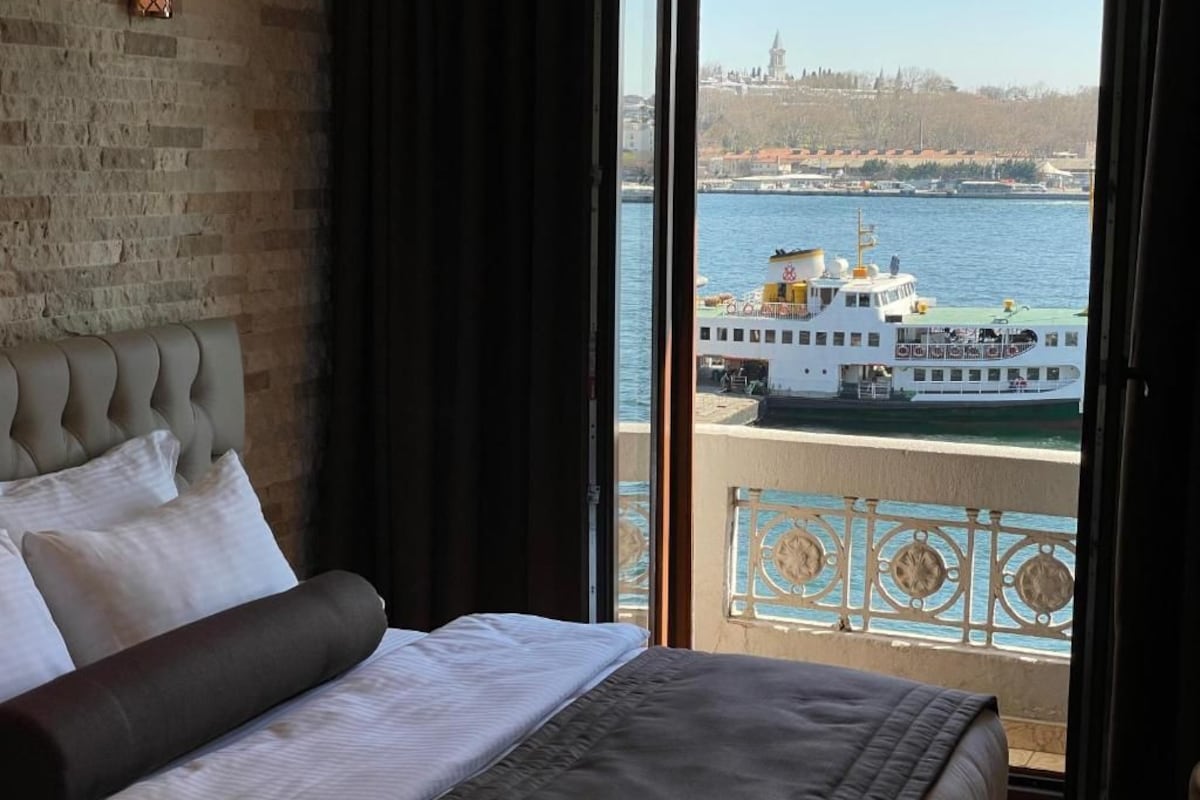 A hotel room with a bed, a view of a river, and a balcony with a railing.