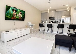 A modern living room with a flat-screen TV, a white entertainment unit, a dining table, and bar stools. - Beautiful Villa at Championsgate (Four Corners)