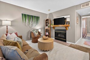 Een woonkamer met een bank, fauteuil, salontafel, open haard, televisie en decoratieve wandversiering.