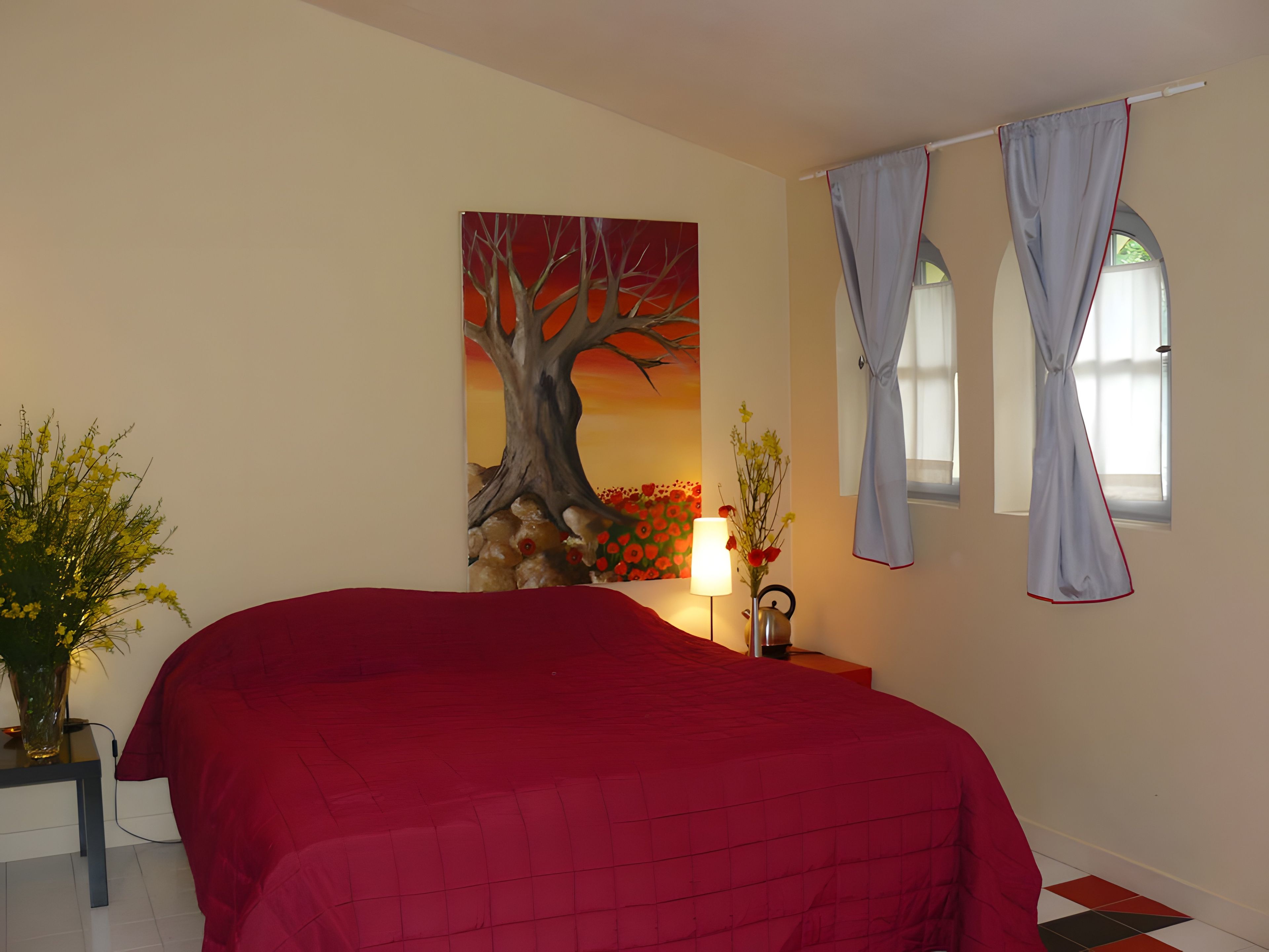 Une chambre à coucher avec un lit, un tableau, un vase de fleurs, une lampe et des rideaux.