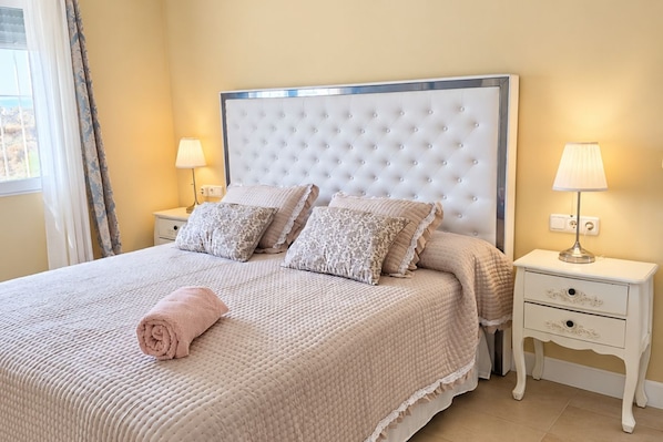 A neatly made bed with a quilted cover, two pillows, and a rolled towel. There are two bedside tables with lamps, one on each side of the bed.