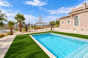 A swimming pool with a pergola, palm trees, and a house with a tiled roof.