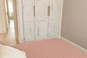 Ein Schlafzimmer mit rosa Bettdecke, weißen Kissen und einem weißen Kleiderschrank mit goldenen Griffen.