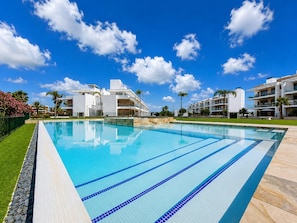 Ein Schwimmbecken mit klarem, blauem Wasser und blauen Bahnen, umgeben von modernen Gebäuden und Palmen unter blauem Himmel mit Wolken.