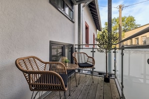 A balcony with wicker chairs and a small table, a potted plant, and a view of a building with red shutters. - City Living—Stylish Living in St. Wendel (Sankt Wendel)