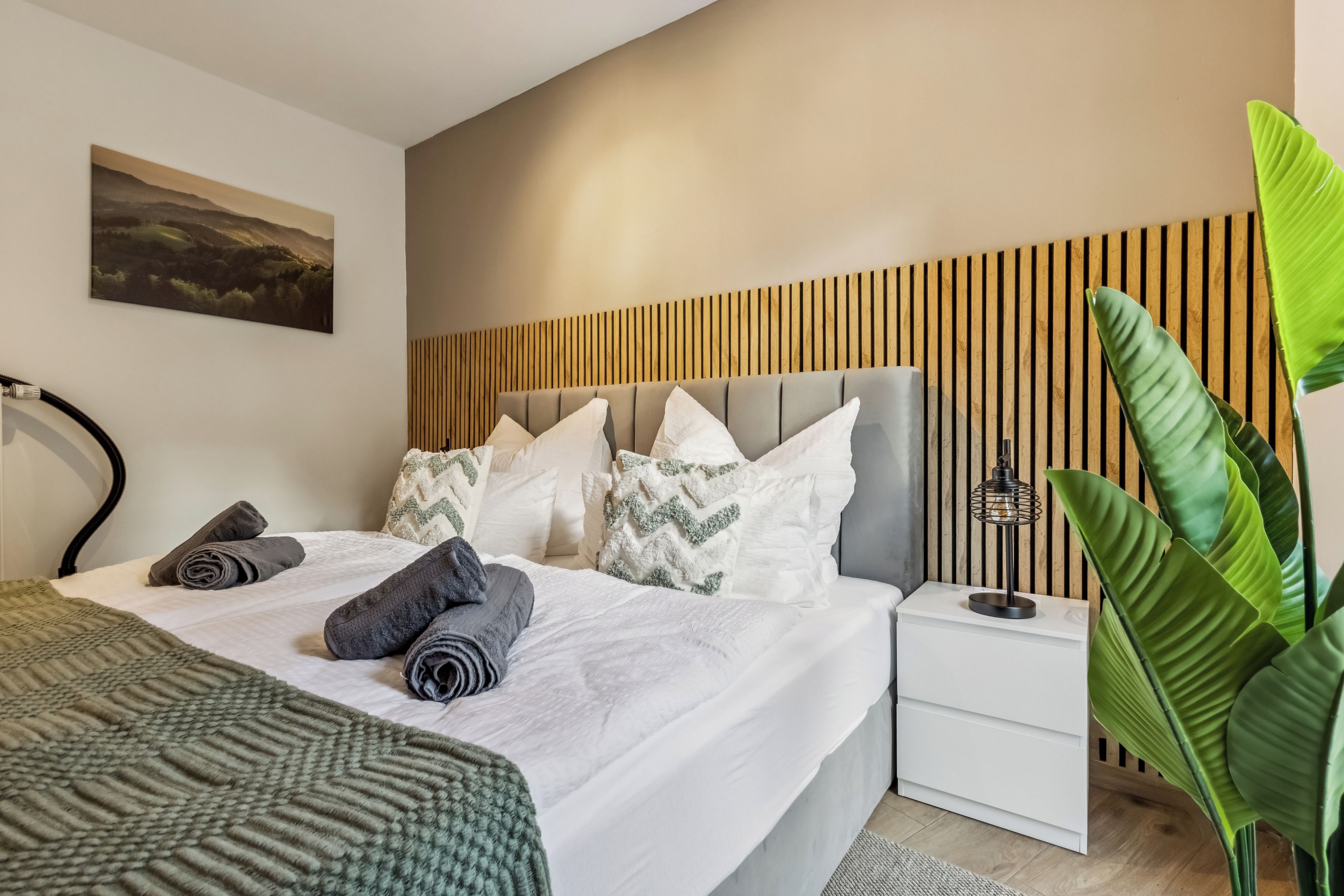 A modern bedroom with a bed, pillows, a nightstand, a lamp, and a large plant.