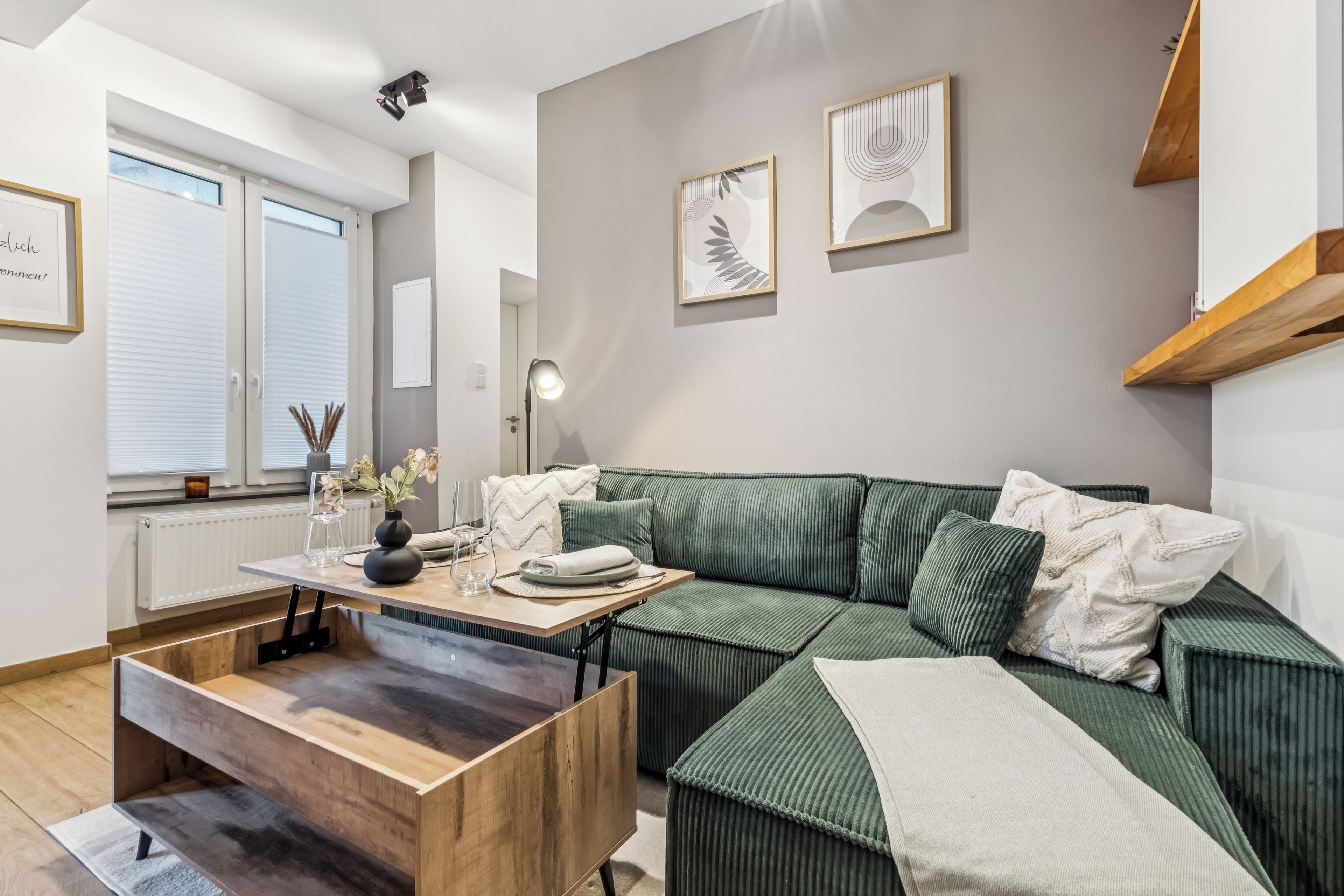 Ein modernes Wohnzimmer mit einem grünen Sofa, einem Holztisch und einem Esstisch mit Stühlen. An der Wand hängen zwei gerahmte Kunstwerke, ein Fenster mit Jalousien und ein Heizkörper.