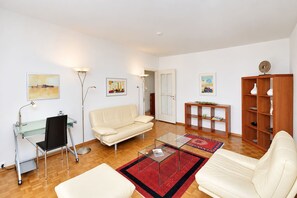Ein modernes Wohnzimmer mit einem Glastisch, einem beigefarbenen Sofa, einem weißen Sessel, einem Schreibtisch mit Stuhl und einem Holzregal.