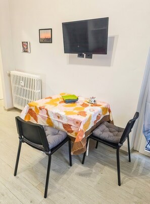 En matplats med ett bord täckt av en blommig duk, två stolar, en tv som är monterad på väggen och ett element.