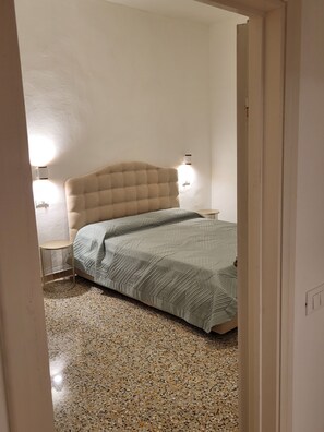 A bedroom with a tufted headboard, a round bedside table, a bed with a patterned bedspread, and a speckled floor.