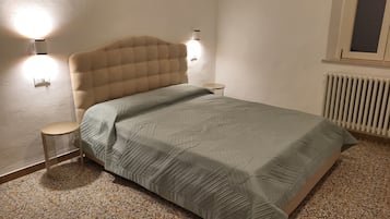Un dormitorio con cabecera capitonada, cama con colcha estampada, dos lámparas de noche, una mesita redonda y un radiador.