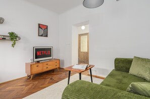 Una sala de estar con un sofá, una mesa de centro, un televisor que muestra 'NETFLIX', un cuadro en la pared y una puerta que da a otra habitación.
